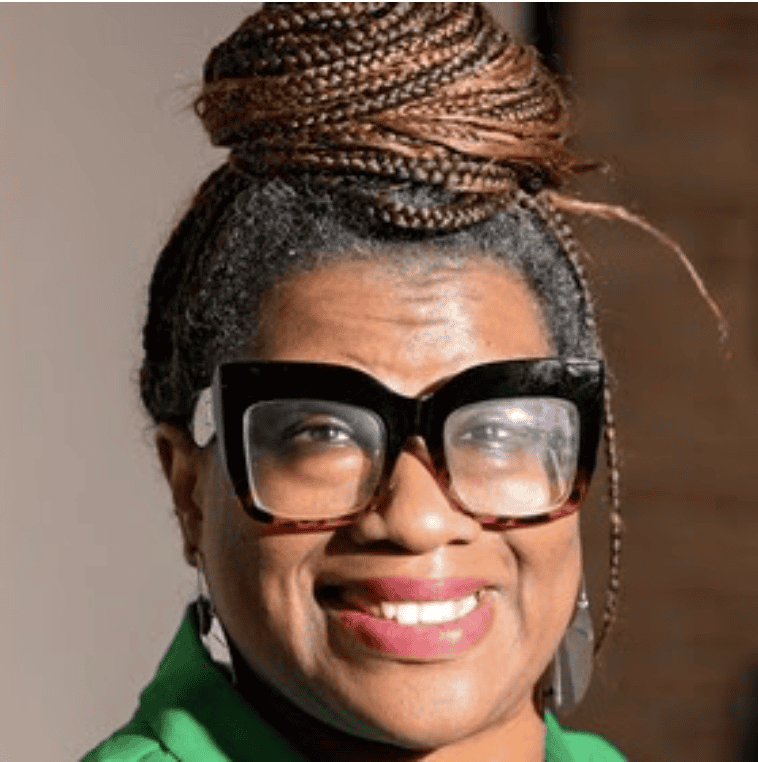 A person with glasses and braided hair poses indoors, wearing a green outfit with bright lighting and a blurred background.