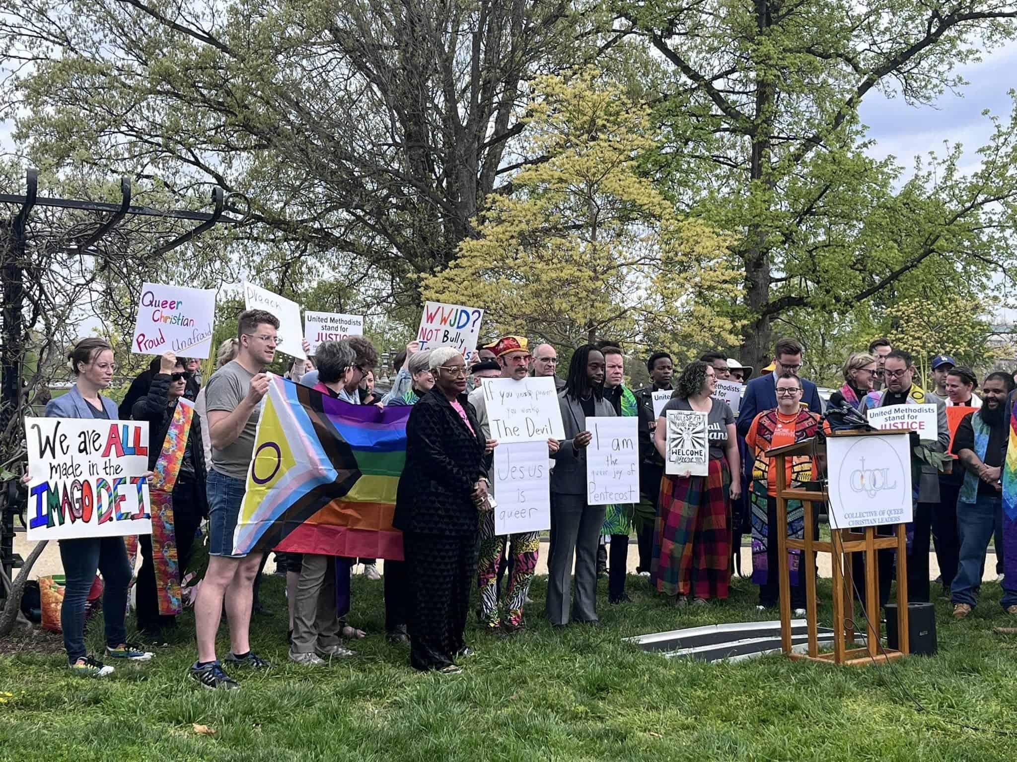 491102875_1093973509440496_1967467485755156280_n A group of people hold signs advocating for LGBTQ+ rights in a park setting with trees in the background.