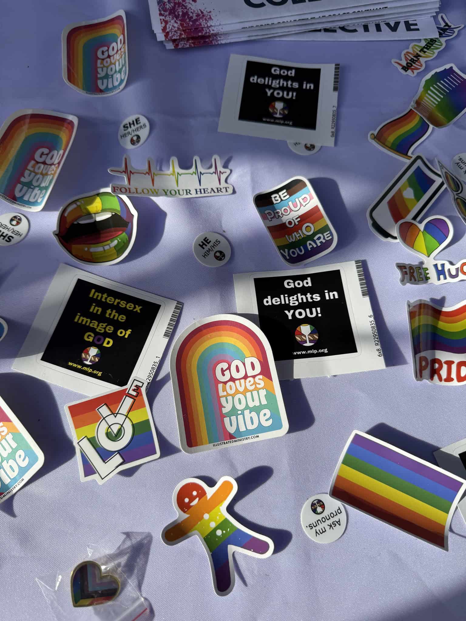 557829832_1232580718913107_6797278852229188311_n Colorful stickers and pins with LGBTQ+ themes, featuring rainbows and affirmations, are spread across a table under bright sunlight.