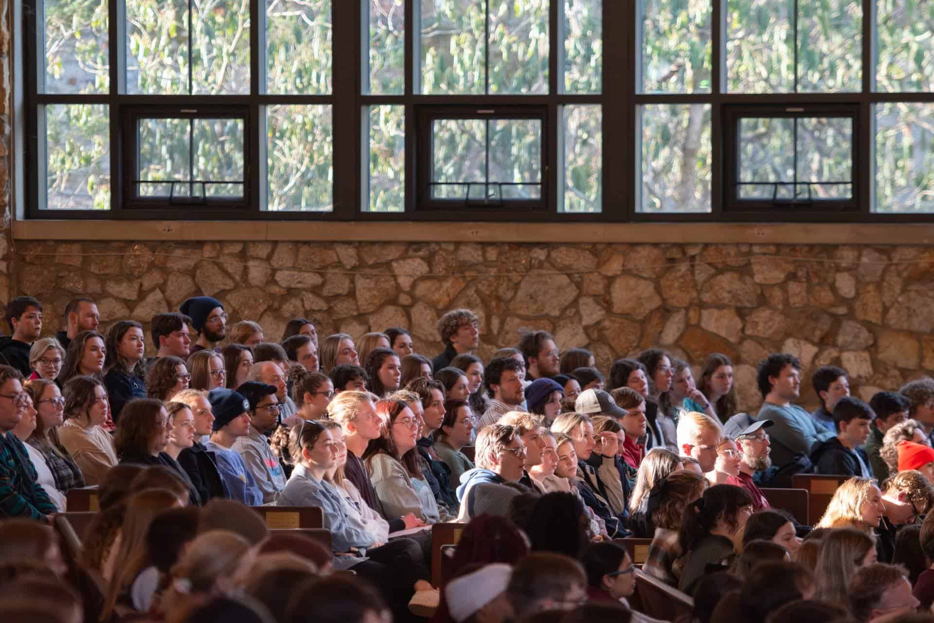 A group of people sitting indoors attentively, in a sunlit room with stone walls and large windows overlooking greenery outside.